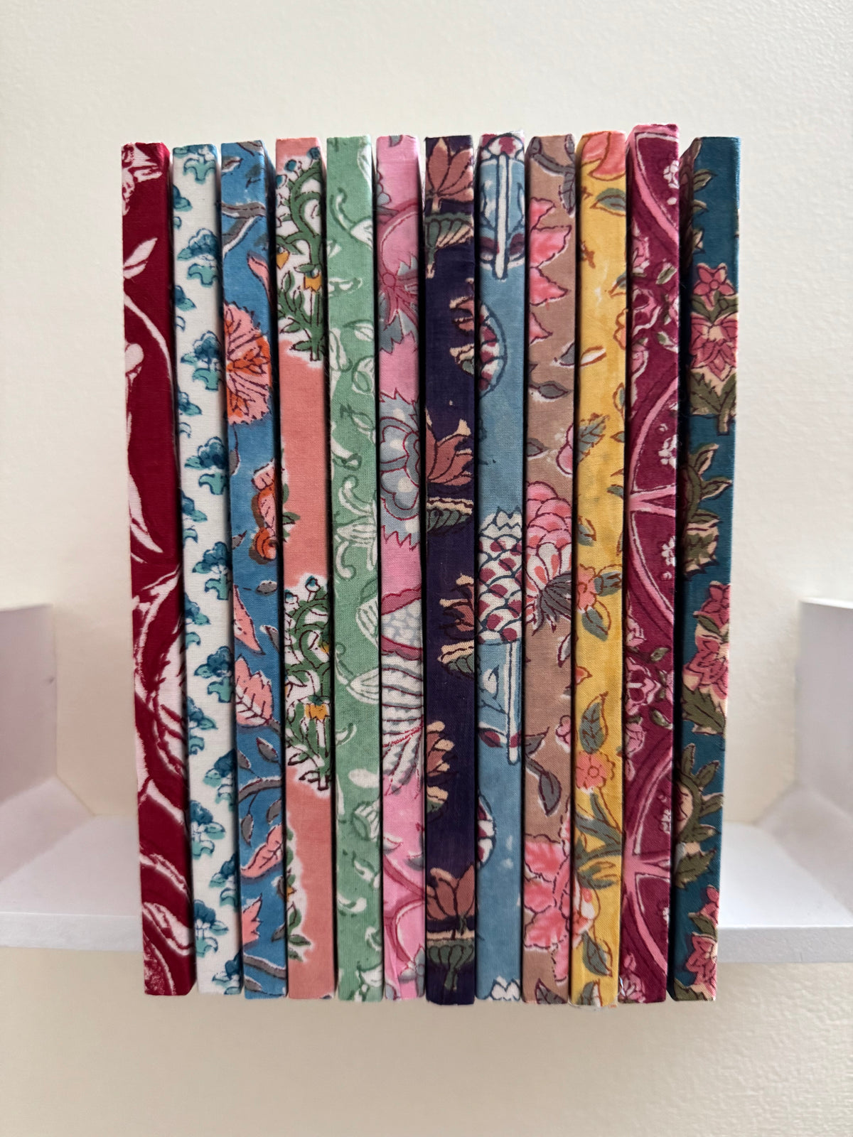 House of Mulmul - Blockprinted Journal Diary Notebook. A set of nine fabric-covered books or book spines standing upright side-by-side, showcasing a variety of colorful floral and botanical patterns in red, blue, green, pink, purple, yellow, and brown tones against a plain off-white background.