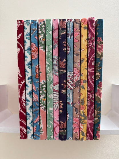 House of Mulmul - Blockprinted Journal Diary Notebook. A set of nine fabric-covered books or book spines standing upright side-by-side, showcasing a variety of colorful floral and botanical patterns in red, blue, green, pink, purple, yellow, and brown tones against a plain off-white background.