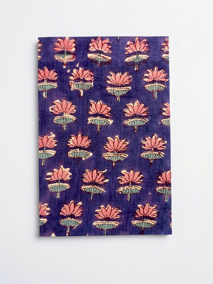 House of Mulmul - Blockprinted Journal Diary Notebook. A rectangular notebook  block-print style lotus flower pattern in pink and green on a deep purple background, displayed flat against a light gray wall.