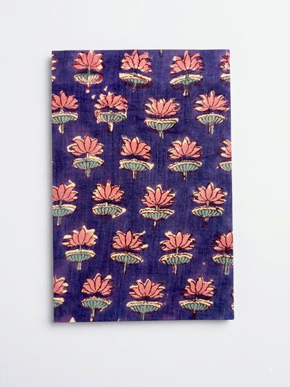 House of Mulmul - Blockprinted Journal Diary Notebook. A rectangular notebook  block-print style lotus flower pattern in pink and green on a deep purple background, displayed flat against a light gray wall.