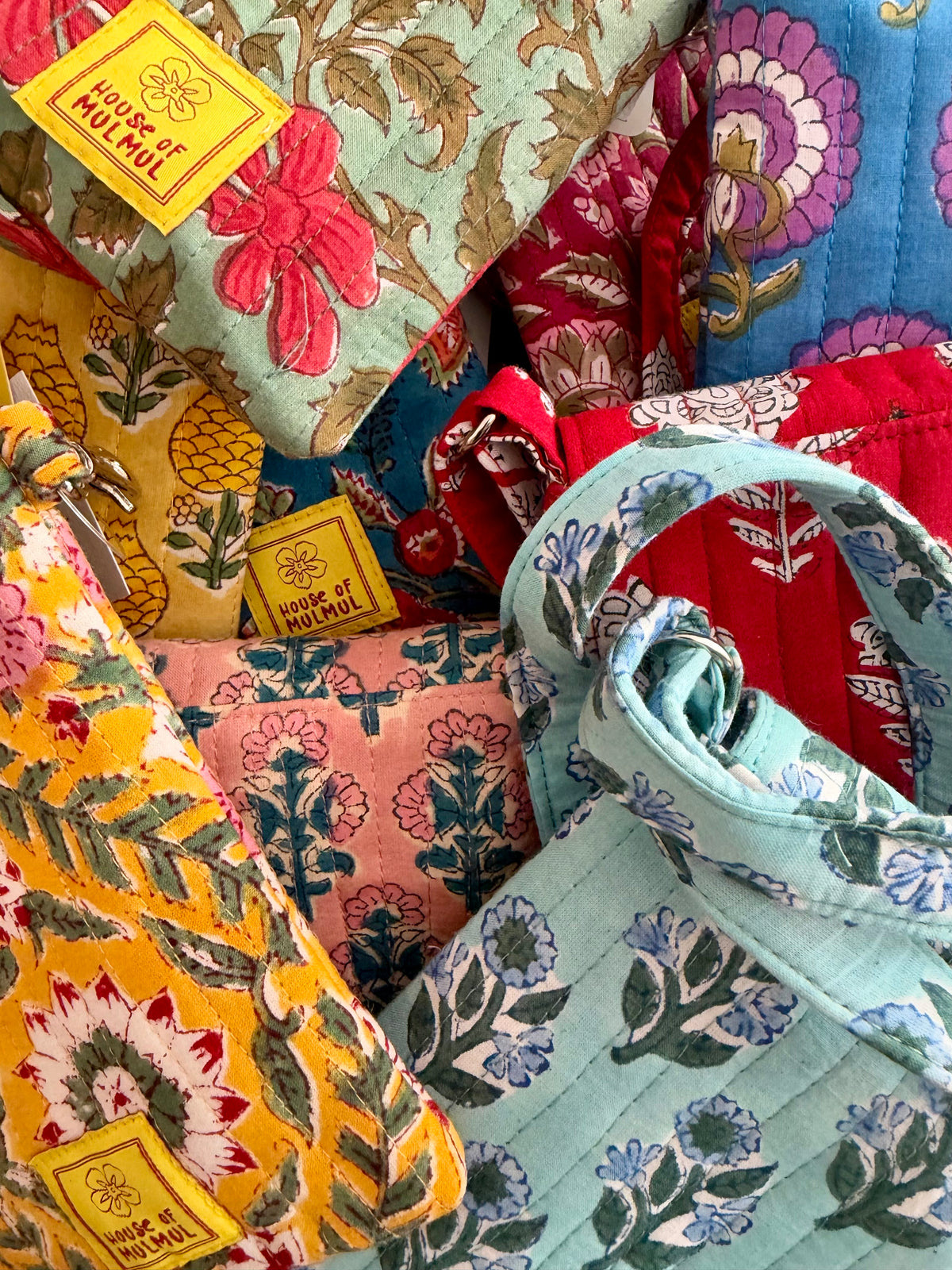 House of Mulmul - Blockprinted Mobile Sling Bag. A close-up of several colorful quilted fabric pouches and small bags with floral and botanical block-print patterns, each showing a yellow 'HOUSE OF MULMUL' label. The collection includes light green, yellow, pink, red, and blue patterns.