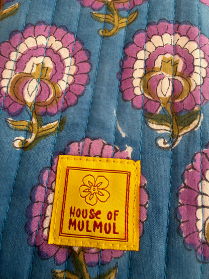House of Mulmul - Blockprinted Mobile Sling Bag. A close-up of a quilted fabric featuring a blue base with a purple, white, and gold floral pattern, finished with a yellow sewn-on label that reads 'HOUSE OF MULMUL'.