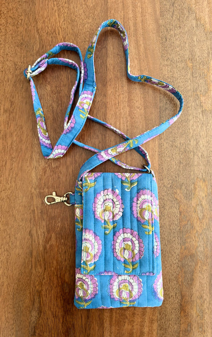 House of Mulmul - Blockprinted Mobile Sling Bag. A small quilted mobile pouch with a matching adjustable shoulder strap, featuring a blue background with pink, purple, and yellow floral motifs, laid flat on a wooden surface.