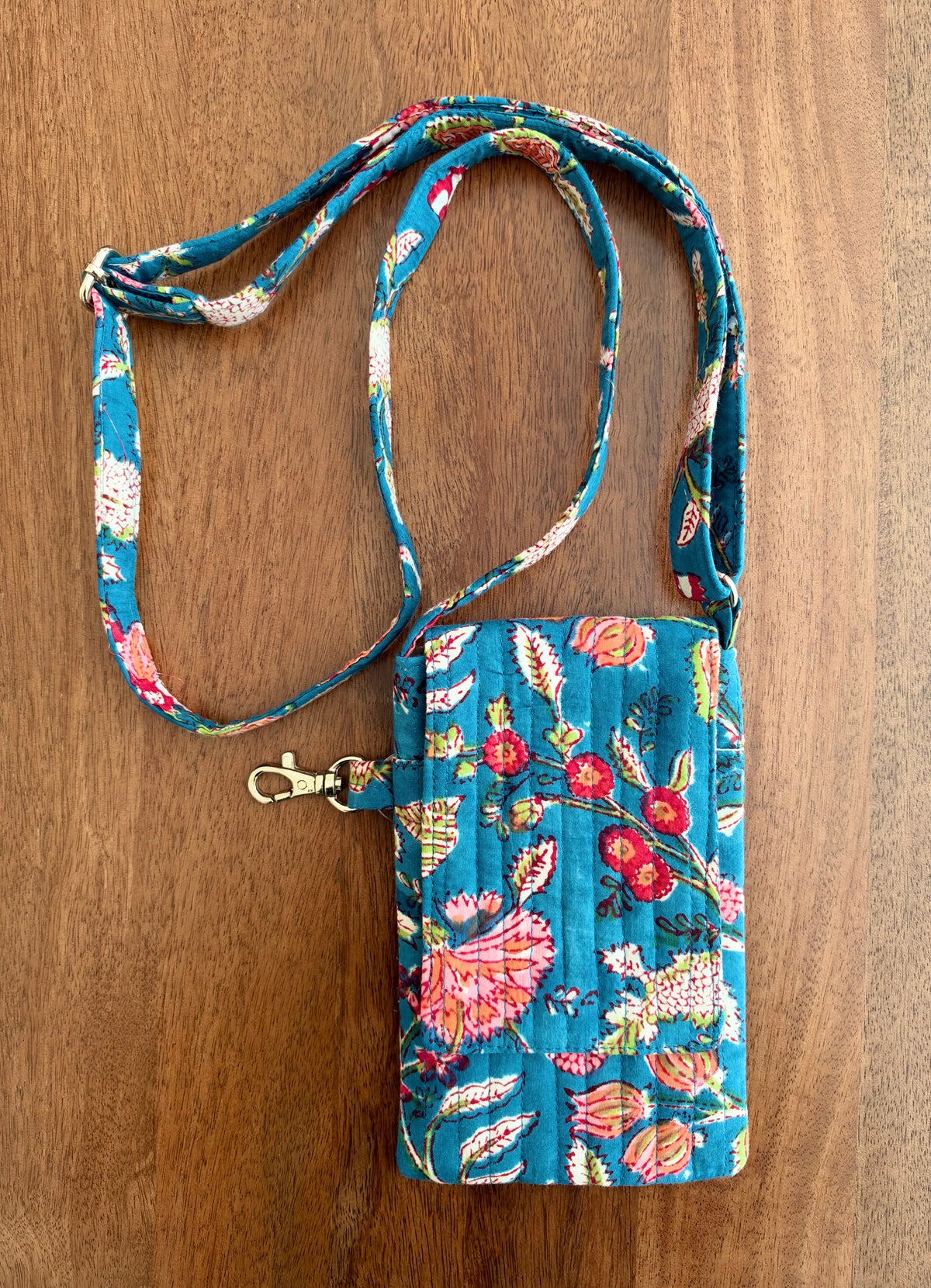 House of Mulmul - Blockprinted Mobile Sling Bag. A small fabric crossbody phone bag with a colorful floral and butterfly print on a teal background, laid flat on a wooden surface. It has a matching fabric strap with a gold-tone clasp.
