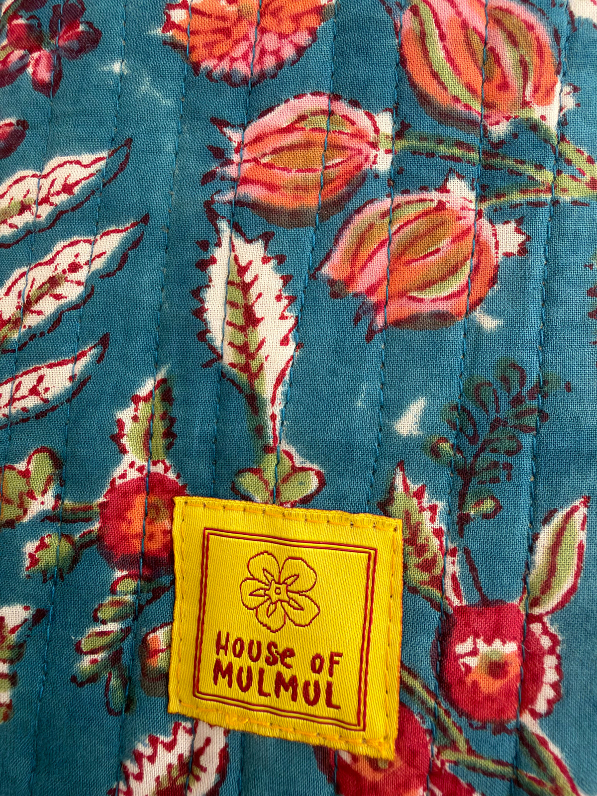 House of Mulmul - Blockprinted Mobile Sling Bag. Close-up of a quilted textile with a teal blue floral pattern in red, pink, and green, featuring a yellow sewn-in label that reads 'House of Mulmul'.