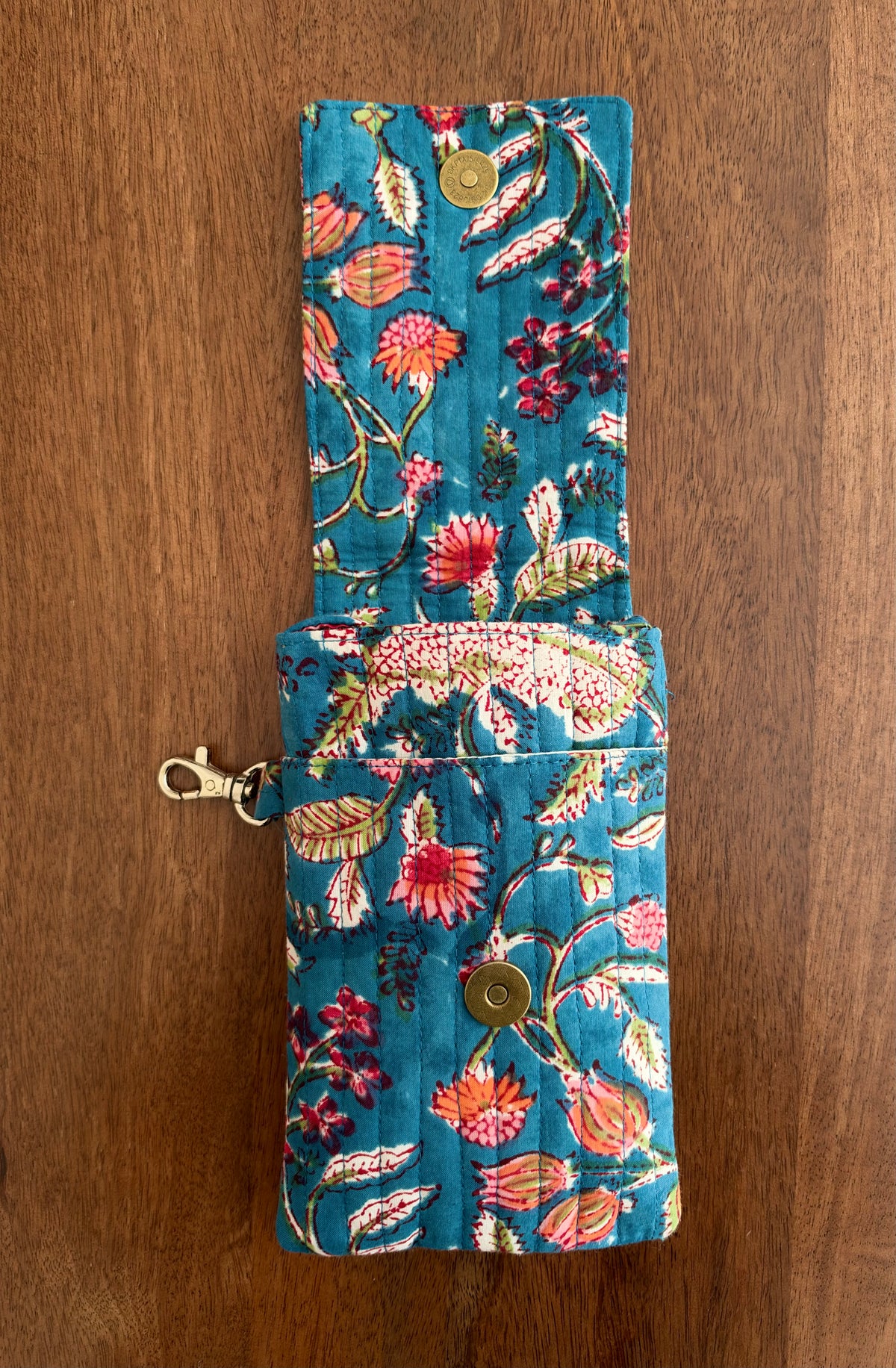 House of Mulmul - Blockprinted Mobile Sling Bag. A fabric pouch with a vibrant floral print on a teal blue background, featuring brass snap buttons and a gold-tone lobster clasp, laid flat on a wooden surface.