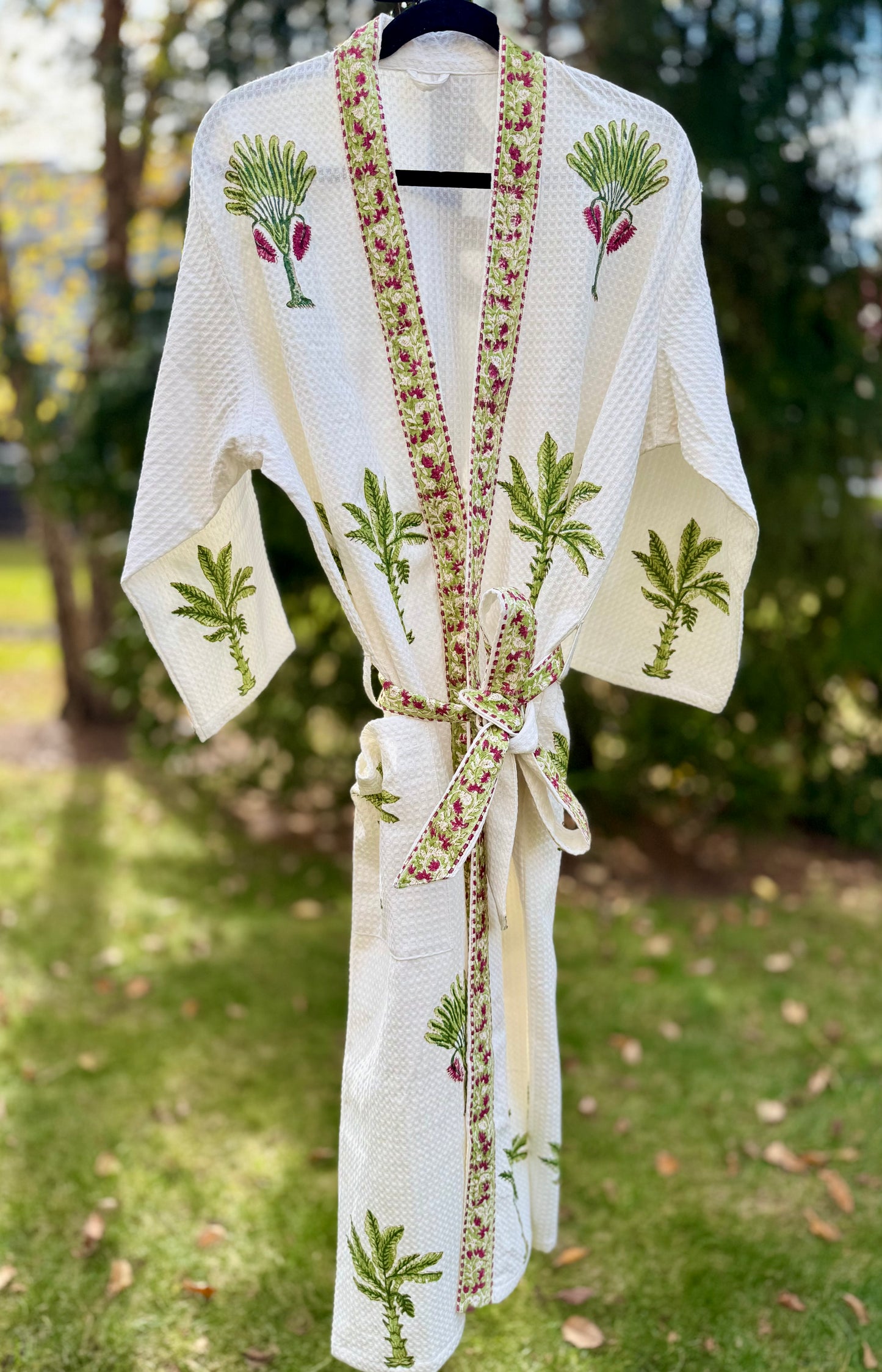 House of Mulmul - Cotton Bath robe. White embroidered robe with green and red patterns on a hanger outdoors.