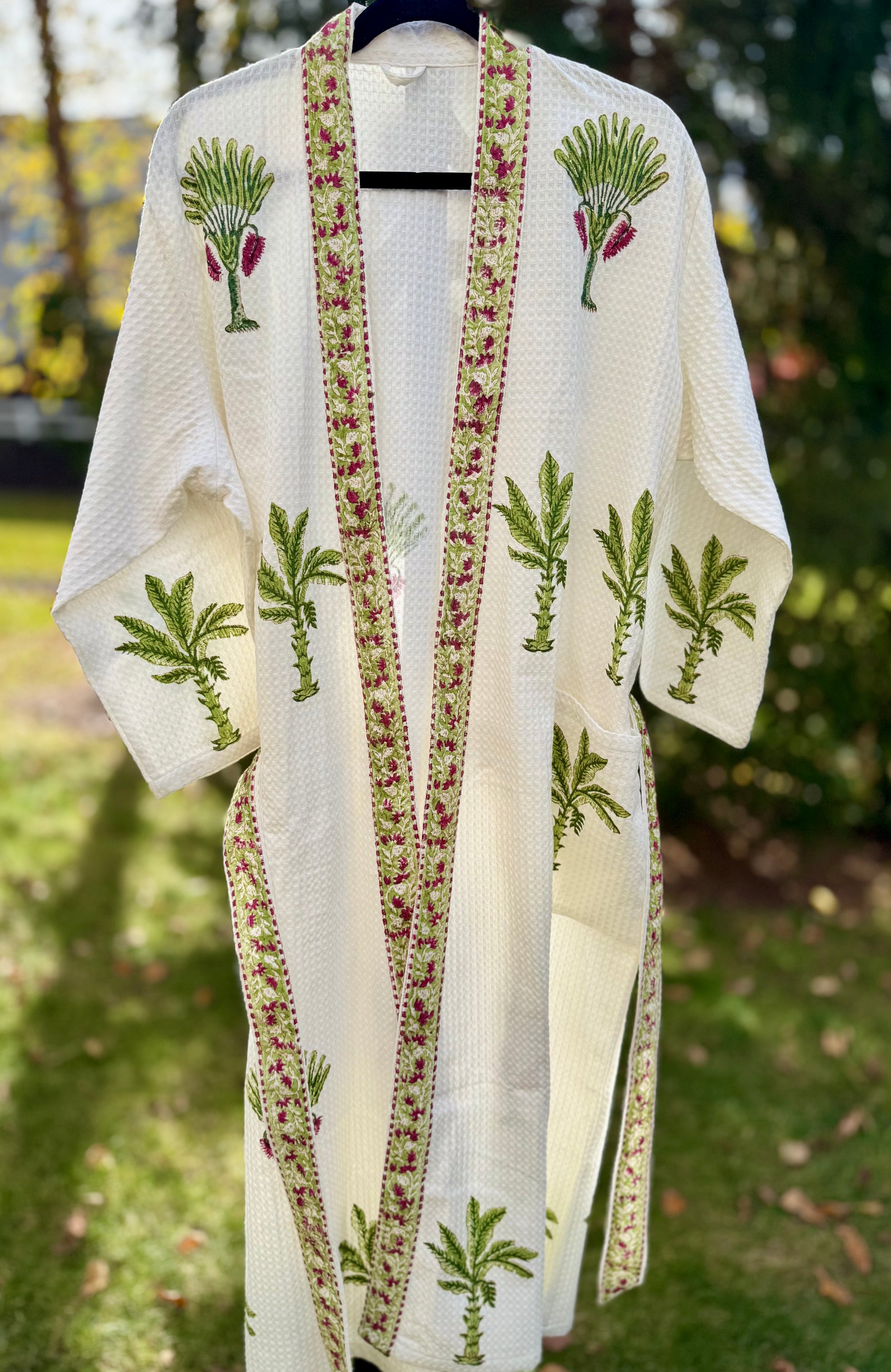 House of Mulmul - Cotton Bath robe. White embroidered robe with green and red patterns on a hanger outdoors.