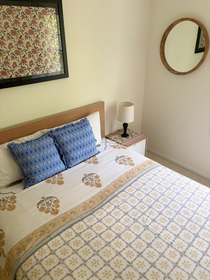 House of Mulmul - Handblock printed cotton summer quilt. Cotton coverlet. Cotton dohar. Muslin blanket. Indian summer quilt. Indian dohar. A neatly made bed in a bedroom setting, featuring a white and beige floral patterned quilt, two blue patterned accent pillows, a wooden headboard, a bedside table with a lamp, and a round wall mirror on the wall.