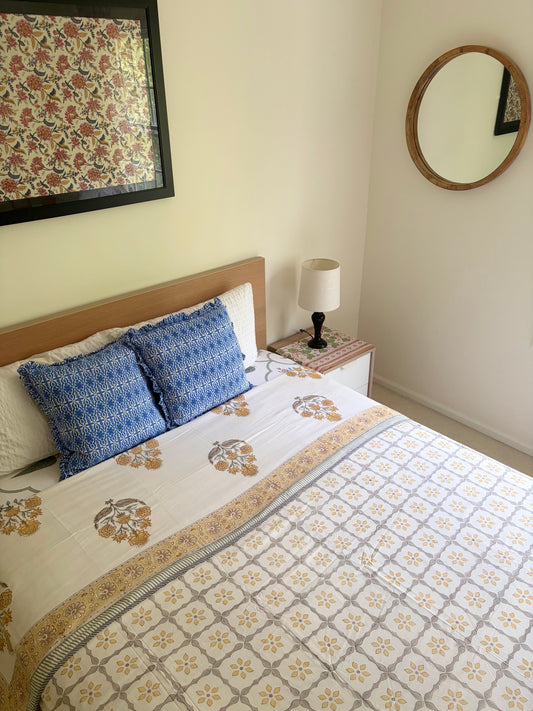 House of Mulmul - Handblock printed cotton summer quilt. Cotton coverlet. Cotton dohar. Muslin blanket. Indian summer quilt. Indian dohar. A neatly made bed in a bedroom setting, featuring a white and beige floral patterned quilt, two blue patterned accent pillows, a wooden headboard, a bedside table with a lamp, and a round wall mirror on the wall.