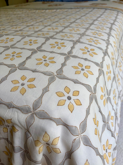 House of Mulmul - Handblock printed cotton summer quilt. Cotton coverlet. Cotton dohar. Muslin blanket. Indian summer quilt. Indian dohar. A close-up of a patterned quilt or comforter with a repeating geometric trellis design in gray and yellow on an off-white background.