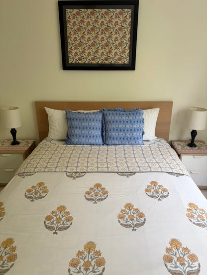 House of Mulmul - Handblock printed cotton summer quilt. Cotton coverlet. Cotton dohar. Muslin blanket. Indian summer quilt. Indian dohar. A neatly made bed in a bedroom setting, featuring a white block-print cotton sheet set with mustard yellow and gray floral motifs, two blue patterned accent pillows, white pillows, and a light wood headboard. A matching framed textile print hangs on the wall above the bed.
