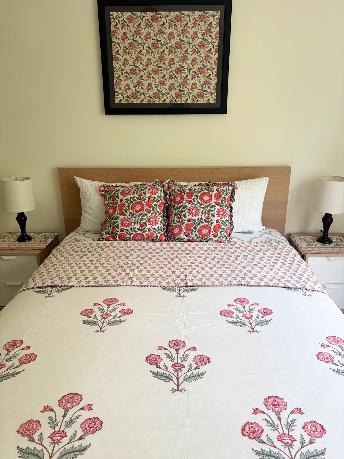 House of Mulmul - Handblock printed cotton summer quilt. Cotton coverlet. Cotton dohar. Muslin blanket. Indian summer quilt. Indian dohar. A neatly made bed in a bedroom setting, featuring a white quilted cover with a pink and green floral pattern, matching patterned pillows, a light wood headboard, bedside tables with lamps, and a framed floral artwork on the wall.