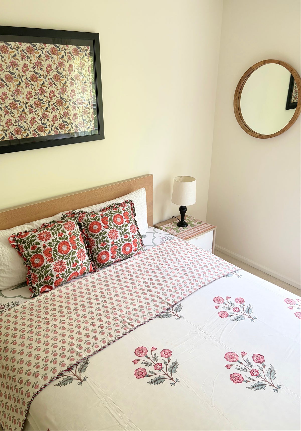 House of Mulmul - Handblock printed cotton summer quilt. Cotton coverlet. Cotton dohar. Muslin blanket. Indian summer quilt. Indian dohar. A styled bedroom scene featuring a bed dressed in a cream-colored floral block-print cotton sheet and matching decorative pillows with red and green floral motifs. A matching framed print hangs on the wall above the headboard, and a small bedside table with a lamp is visible.