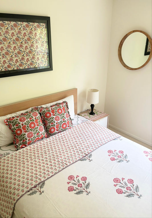 House of Mulmul - Handblock printed cotton summer quilt. Cotton coverlet. Cotton dohar. Muslin blanket. Indian summer quilt. Indian dohar. A styled bedroom scene featuring a bed dressed in a cream-colored floral block-print cotton sheet and matching decorative pillows with red and green floral motifs. A matching framed print hangs on the wall above the headboard, and a small bedside table with a lamp is visible.