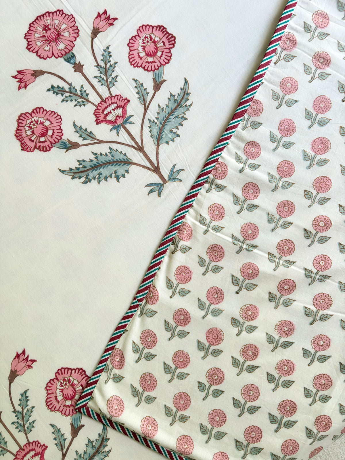 House of Mulmul - Handblock printed cotton summer quilt. Cotton coverlet. Cotton dohar. Muslin blanket. Indian summer quilt. Indian dohar. A close-up of a cream-colored fabric with pink floral block prints and green leaves, folded diagonally to show a striped red, green, and blue border.