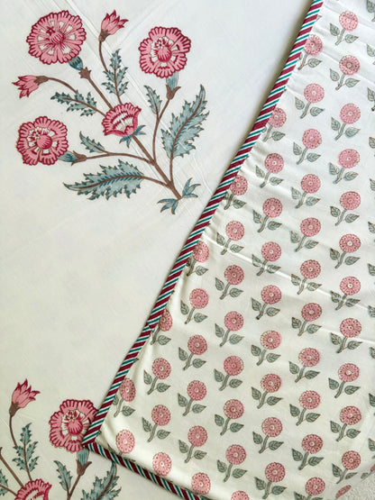 House of Mulmul - Handblock printed cotton summer quilt. Cotton coverlet. Cotton dohar. Muslin blanket. Indian summer quilt. Indian dohar. A close-up of a cream-colored fabric with pink floral block prints and green leaves, folded diagonally to show a striped red, green, and blue border.