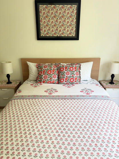 House of Mulmul - Handblock printed cotton summer quilt. Cotton coverlet. Cotton dohar. Muslin blanket. Indian summer quilt. Indian dohar. A neatly made bed in a bedroom setting, featuring a white cotton bedspread and matching pillow shams with a pink and green floral block print. Two additional solid white pillows are placed behind them, flanked by two white nightstands with black table lamps.
