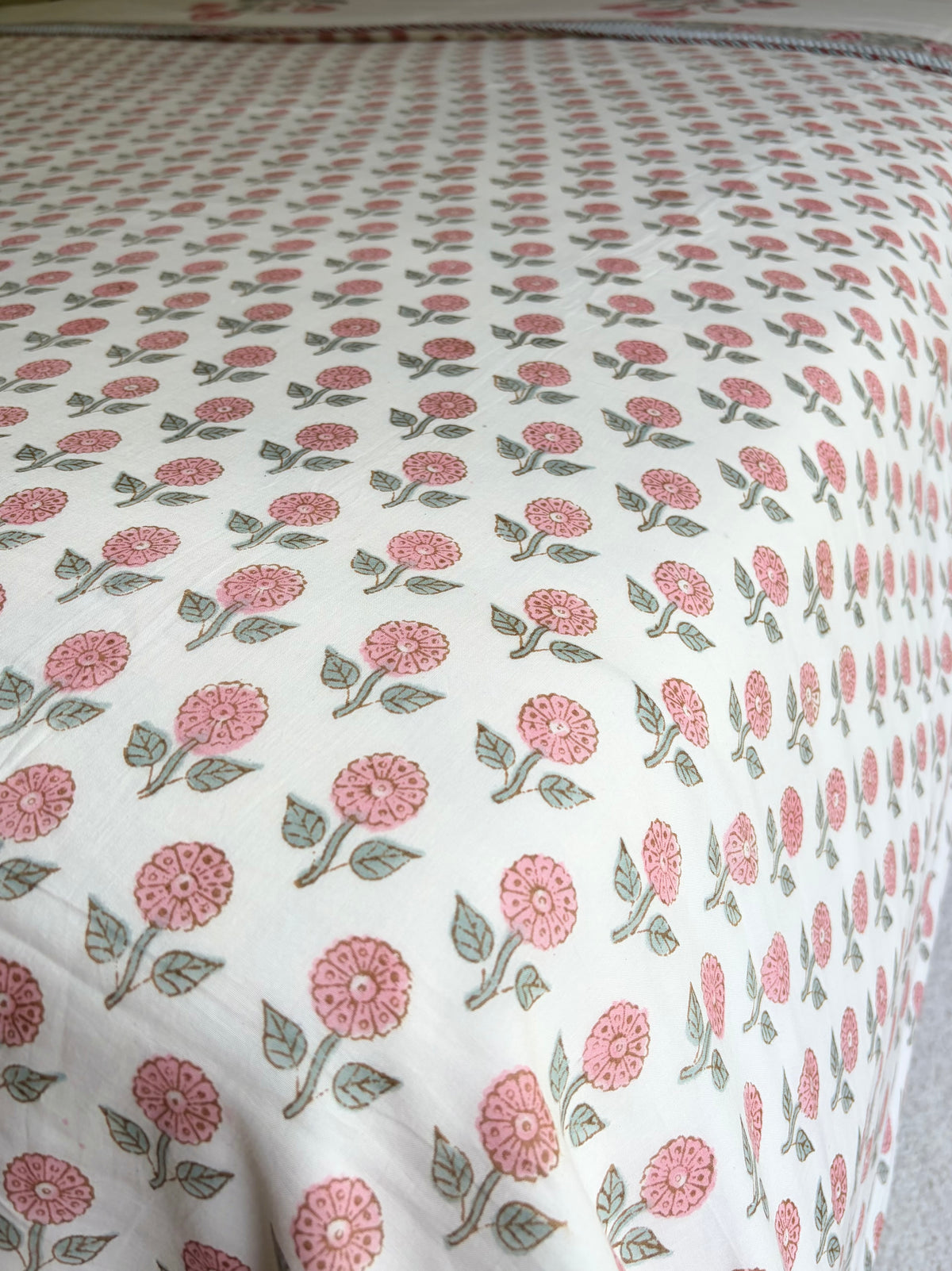 House of Mulmul - Handblock printed cotton summer quilt. Cotton coverlet. Cotton dohar. Muslin blanket. Indian summer quilt. Indian dohar. A close-up of a white fabric, likely a quilt or comforter, featuring a repeating block-print style pattern of pink flowers and green leaves.