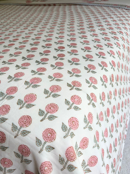 House of Mulmul - Handblock printed cotton summer quilt. Cotton coverlet. Cotton dohar. Muslin blanket. Indian summer quilt. Indian dohar. A close-up of a white fabric, likely a quilt or comforter, featuring a repeating block-print style pattern of pink flowers and green leaves.