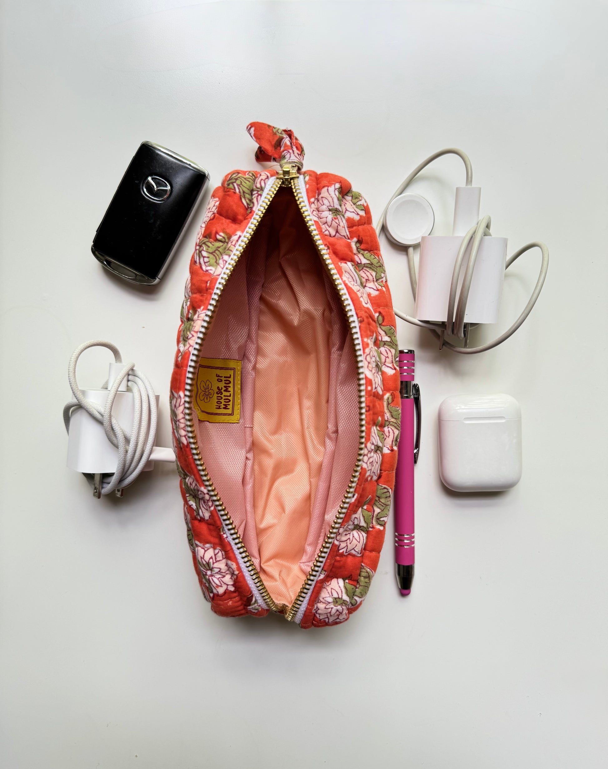 House of Mulmul - Cotton Quilted Makeup Barrel Pouch. An open floral zippered pouch with a pink satin lining, surrounded by travel accessories on a white background, including a black power bank, white chargers, AirPods case, and a pink pen.