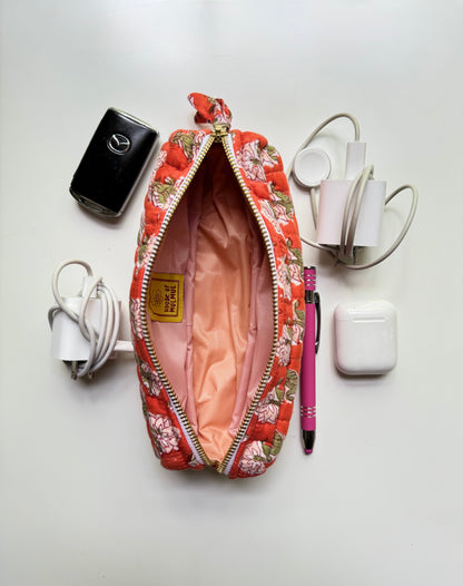 House of Mulmul - Cotton Quilted Makeup Barrel Pouch. An open floral zippered pouch with a pink satin lining, surrounded by travel accessories on a white background, including a black power bank, white chargers, AirPods case, and a pink pen.