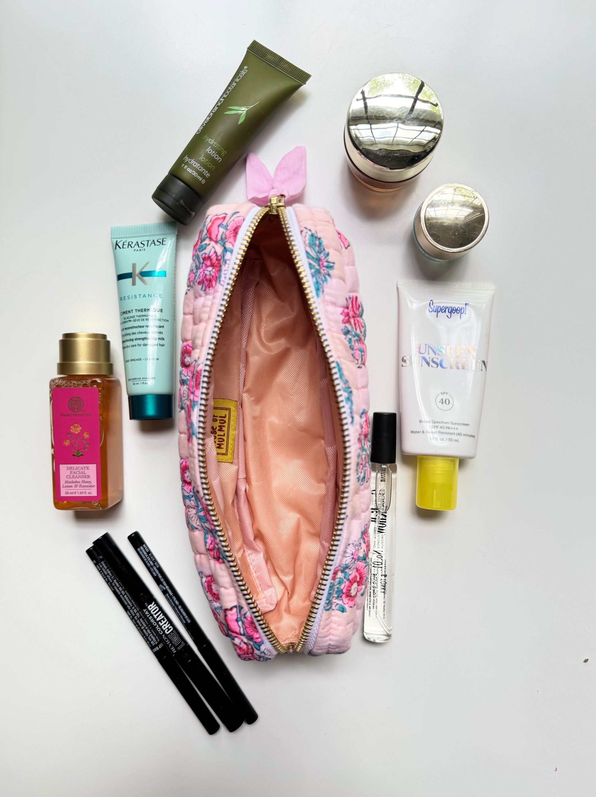 House of Mulmul - Cotton Quilted Makeup Barrel Pouch. A pink floral zippered cosmetic pouch with a gold zipper and a small pink bow on top, shown open with various beauty products arranged around it on a white background. Visible products include a Kiehl green tube, a Kiehl teal tube, a Kiehl white tube, a Kiehl gold bottle, two black pencils, and a small clear spray bottle.