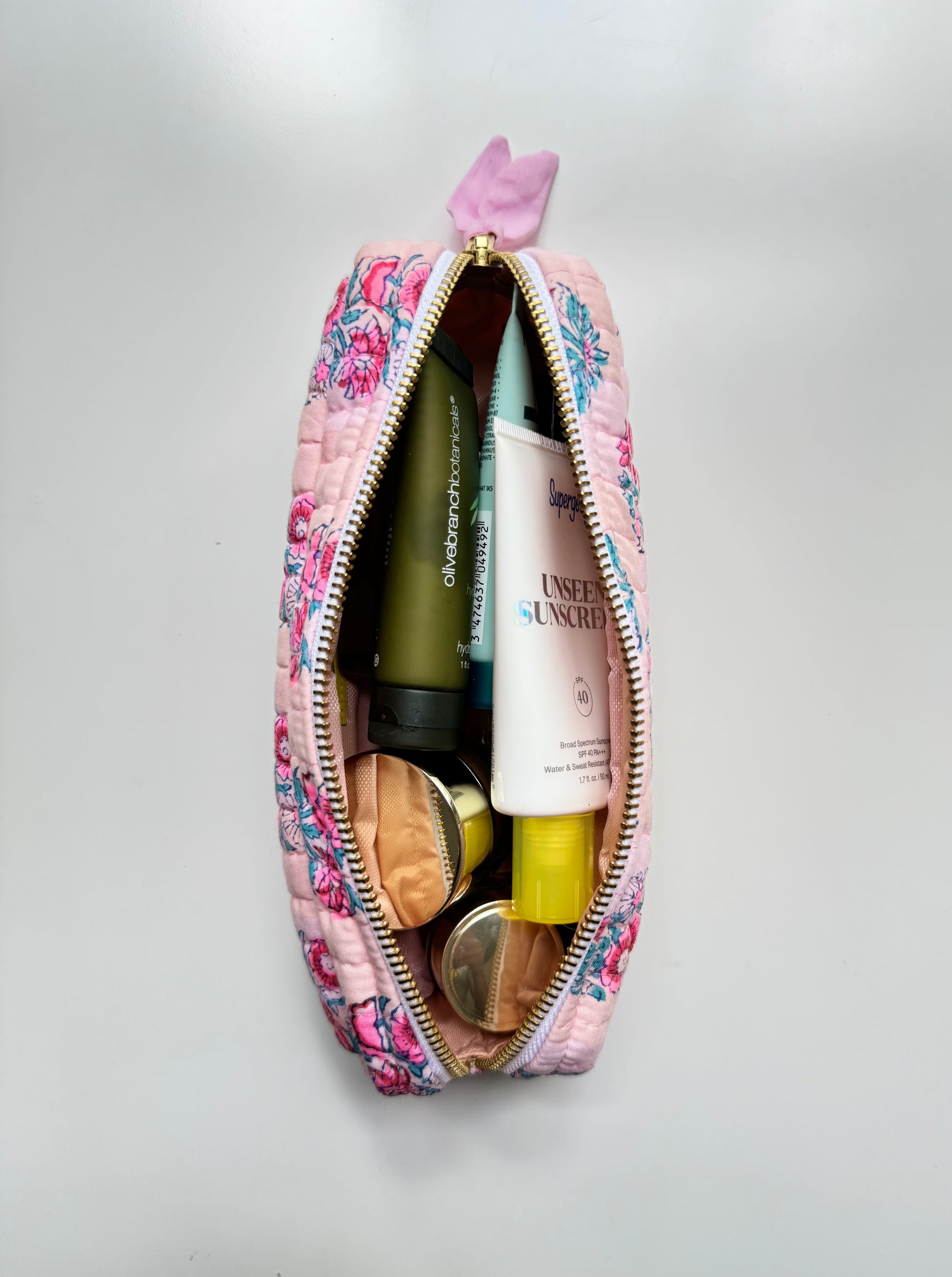 House of Mulmul - Cotton Quilted Makeup Barrel Pouch. A pink quilted cosmetic pouch with a floral print and a small purple bow on the zipper pull, shown open to reveal various makeup and skincare items inside, set against a plain light gray background.
