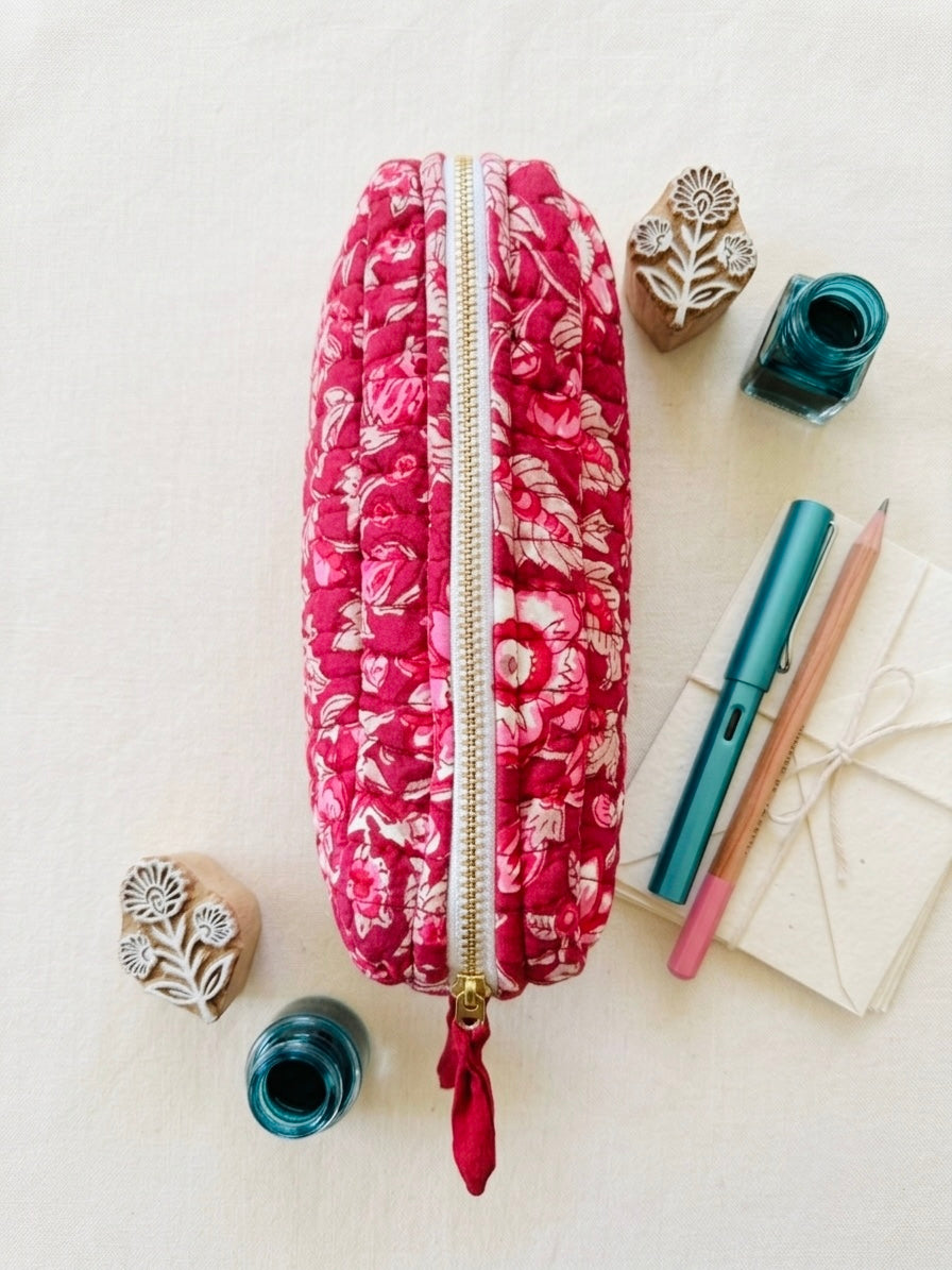House of Mulmul - Cotton Quilted Makeup Barrel Pouch. A pink floral zippered pouch with a red tassel zipper pull, styled in a flat lay with stationery and ink bottles on a light beige textured background.