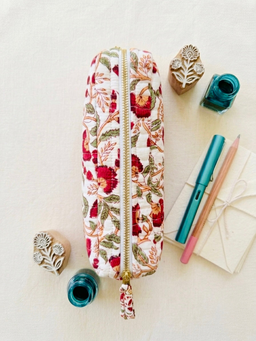 House of Mulmul - Cotton Quilted Makeup Barrel Pouch. A fabric zippered pencil case with a red and green floral print, styled in a flat lay with stationery and rubber stamps on a light cream background.