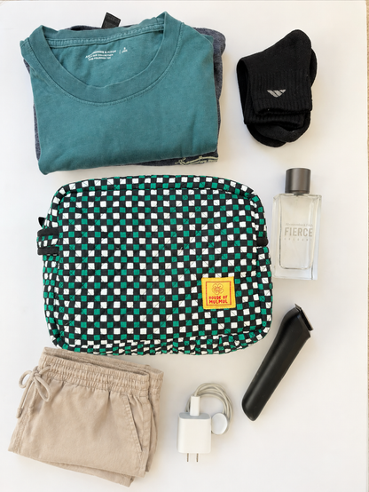House of Mulmul - Handblock Printed Travel Makeup Quilted Pouch set. A flat-lay arrangement of travel items on a white background, featuring a green and white checkered toiletry bag, a teal t-shirt, black socks, beige shorts, a perfume bottle, a black trimmer, and a white phone charger.