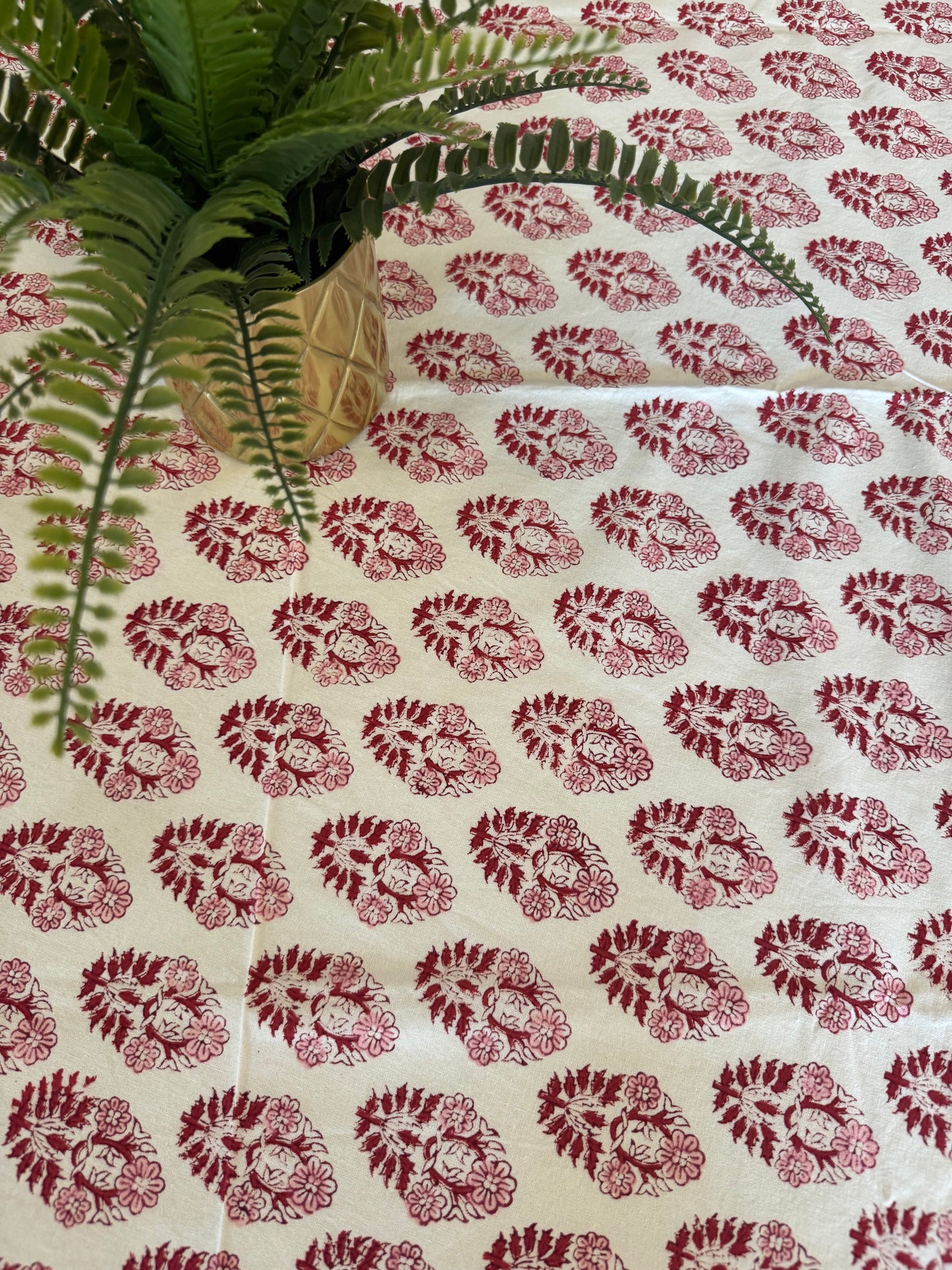 House of Mulmul - Cotton Table Cloth Round. Fabric with red floral pattern on a white background, with green leaves in the foreground.