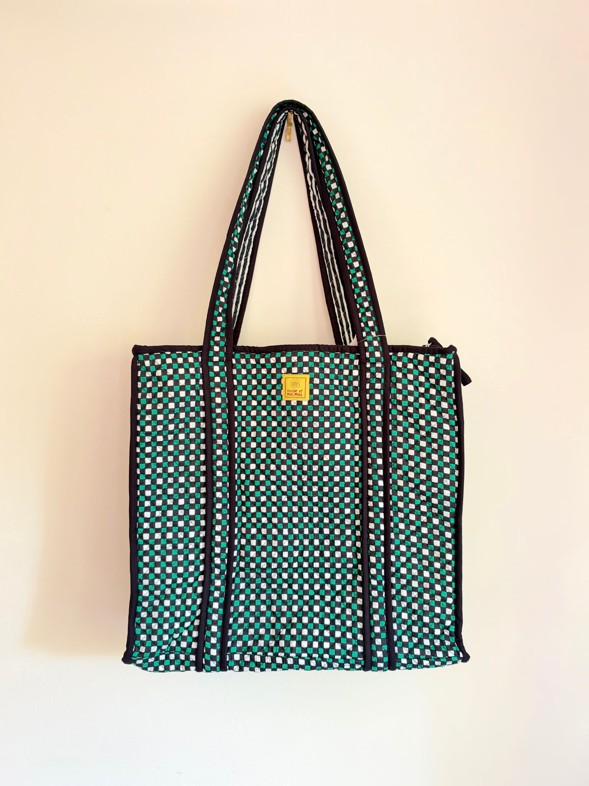 House of Mulmul - Cotton Tote Bags. A green tote bag with a white polka dot pattern, black trim, and two shoulder straps, hanging against a plain off-white wall.