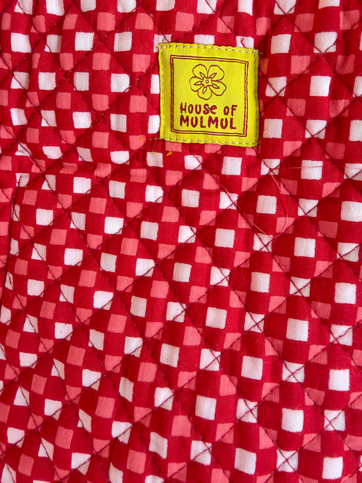 House of Mulmul - Cotton Tote Bags. Close-up of a red and white checkered quilted fabric with a yellow sewn-in label reading 'HOUSE OF MULMUL'.