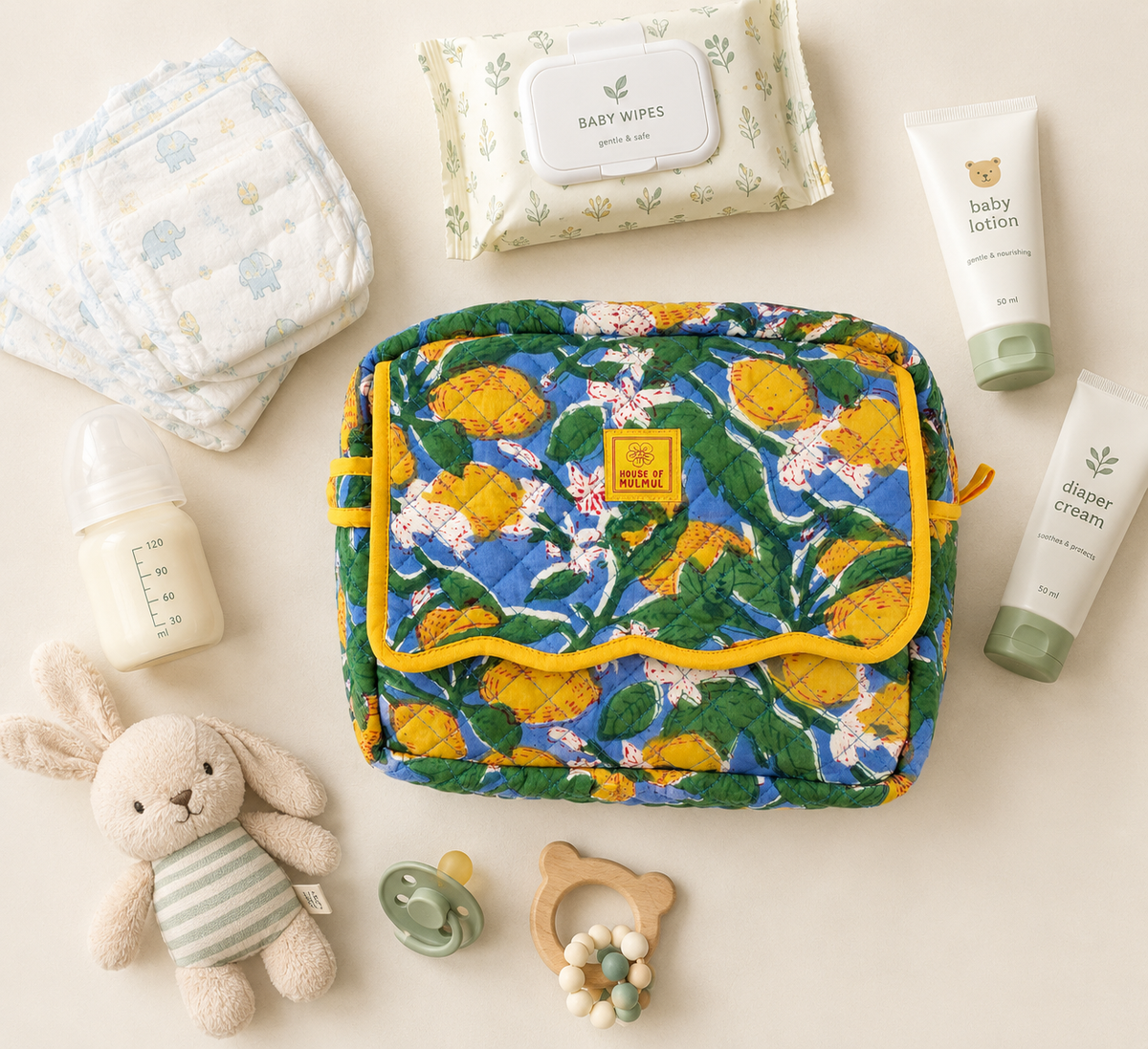 House of Mulmul - Handblock Printed Travel Makeup Quilted Pouch setA quilted baby toiletry bag with a yellow and green lemon pattern and yellow trim, styled in a flat lay with baby items including a diaper, baby wipes, bottle, lotion, diaper cream, pacifier, teething ring, and plush bunny on a cream background.