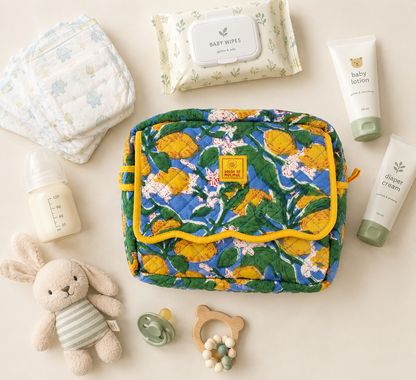 House of Mulmul - Handblock Printed Travel Makeup Quilted Pouch setA quilted baby toiletry bag with a yellow and green lemon pattern and yellow trim, styled in a flat lay with baby items including a diaper, baby wipes, bottle, lotion, diaper cream, pacifier, teething ring, and plush bunny on a cream background.