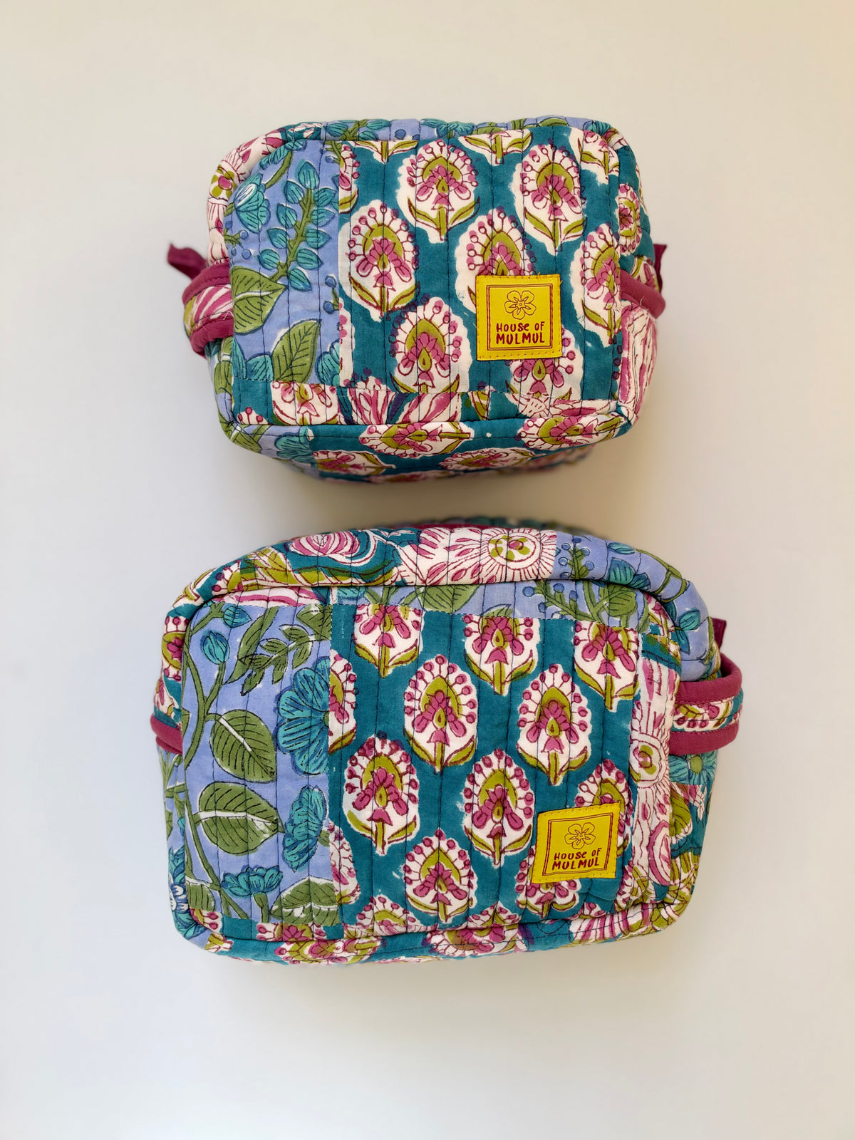 House of Mulmul - Handblock Printed Travel Makeup Quilted Pouch set. Two quilted cosmetic pouches with a colorful floral and botanical print, displayed vertically on a plain white background. Each pouch features a yellow brand label reading 'BAG OF TRICKS' and has a pink strap.