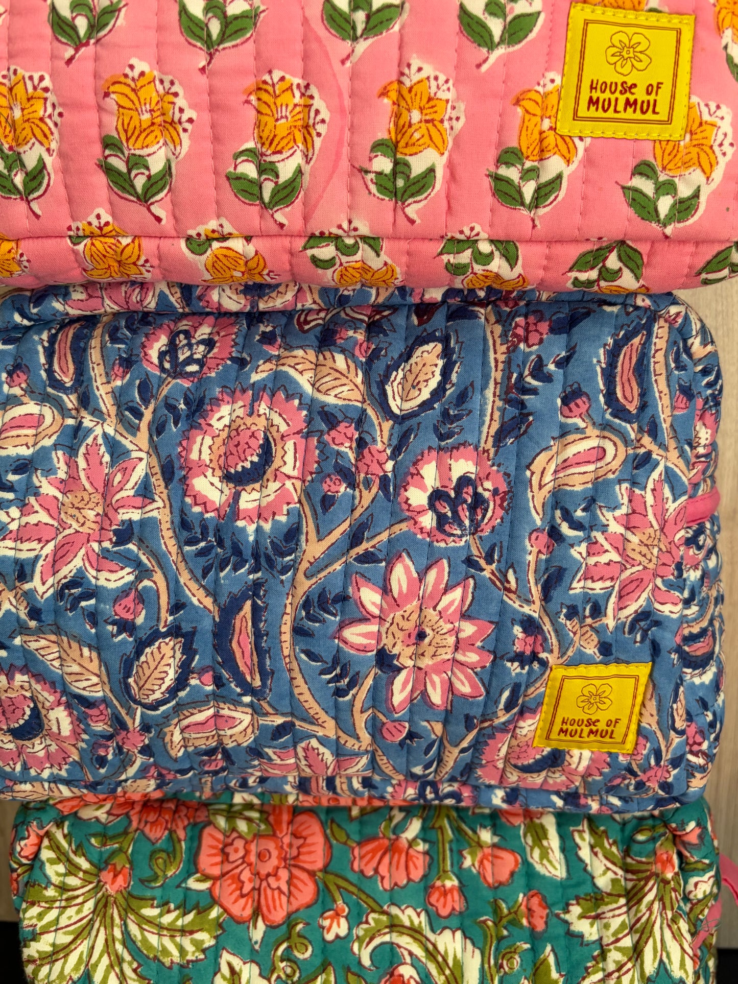 House of Mulmul - Quilted Blockprint Travel Makeup pouch. Three floral-patterned makeup pouches of brand  'House of Mulmul' labels on a pink, blue, and green background.