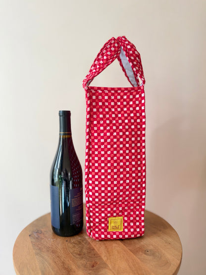 House of Mulmul - Quilted Wine Tote Bag. A red and white checkered fabric wine tote bag with a top handle stands on a round wooden table next to a dark wine bottle against a plain beige wall.
