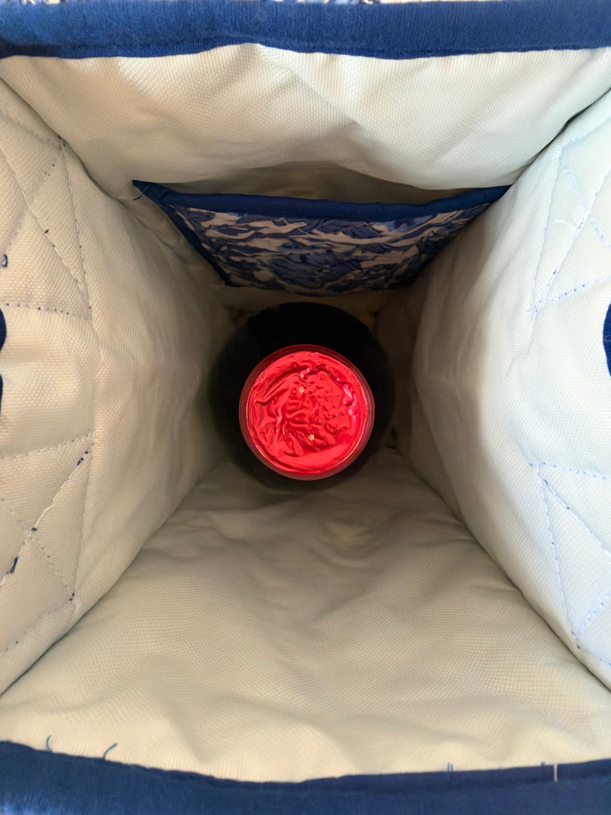 House of Mulmul - Quilted Wine Tote Bag. An interior view looking down into a quilted insulated bag with white fabric lining and blue trim, containing a dark bottle with a bright red cap or label.