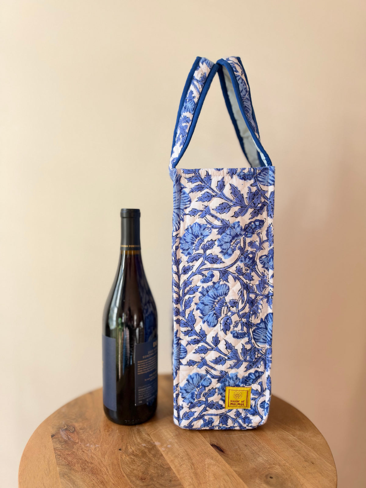 House of Mulmul - Quilted Wine Tote Bag. A blue and white floral patterned wine tote bag with a small yellow label, standing on a wooden stool next to a dark wine bottle against a beige background.