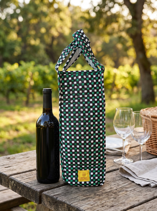 House of Mulmul - Quilted Wine Tote Bag. A green and white checkered wine bottle carrier bag stands on a rustic wooden picnic table outdoors, next to a dark wine bottle, two clear wine glasses, and a wicker basket. The background shows a softly blurred vineyard landscape.