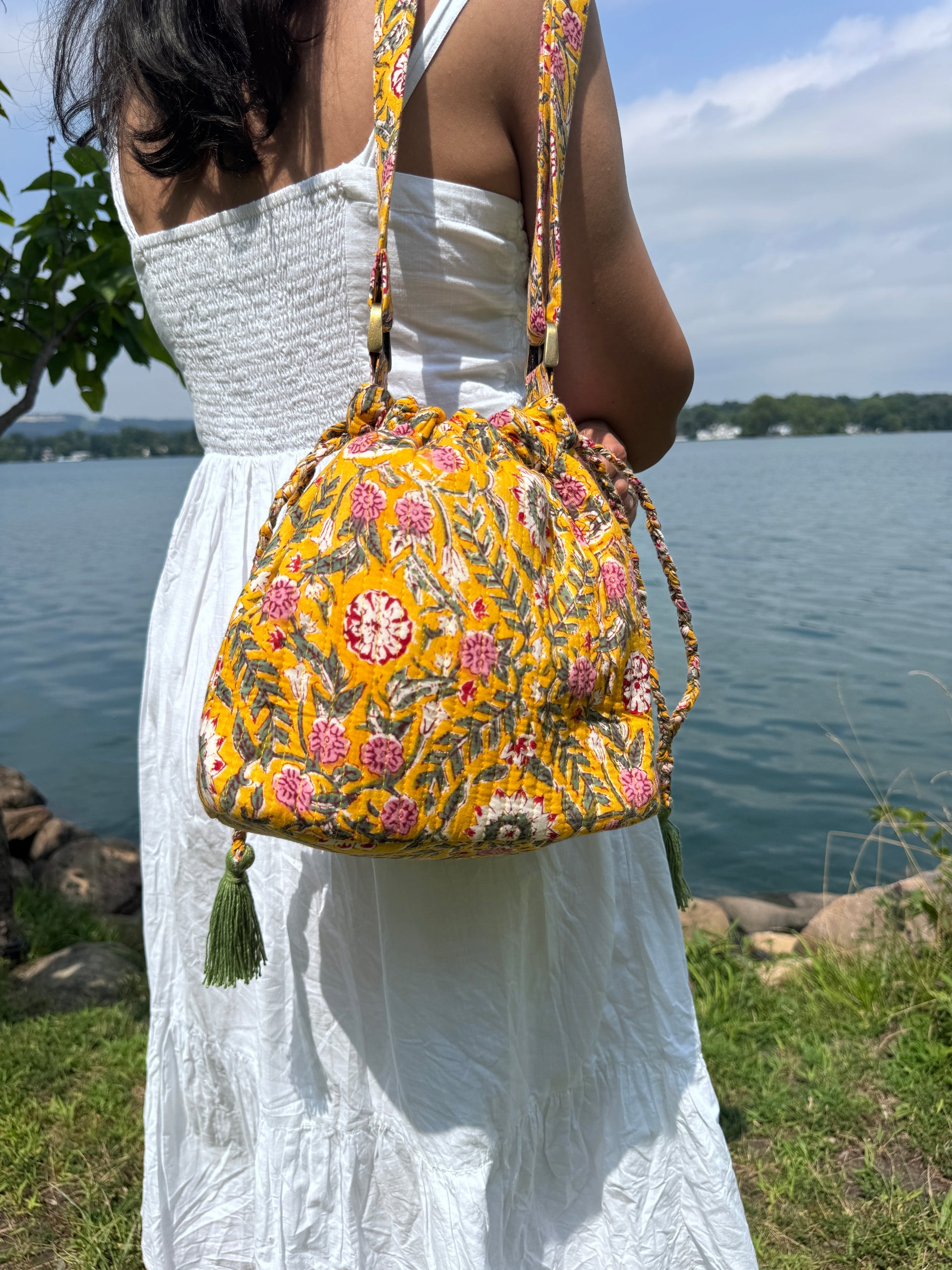 House of Mulmul - Garden Wayfarer Bucket Bag, Cotton bag, Crossbody sling bag. Person holding a yellow floral bag outdoors with a white tarp and blue sky in the background