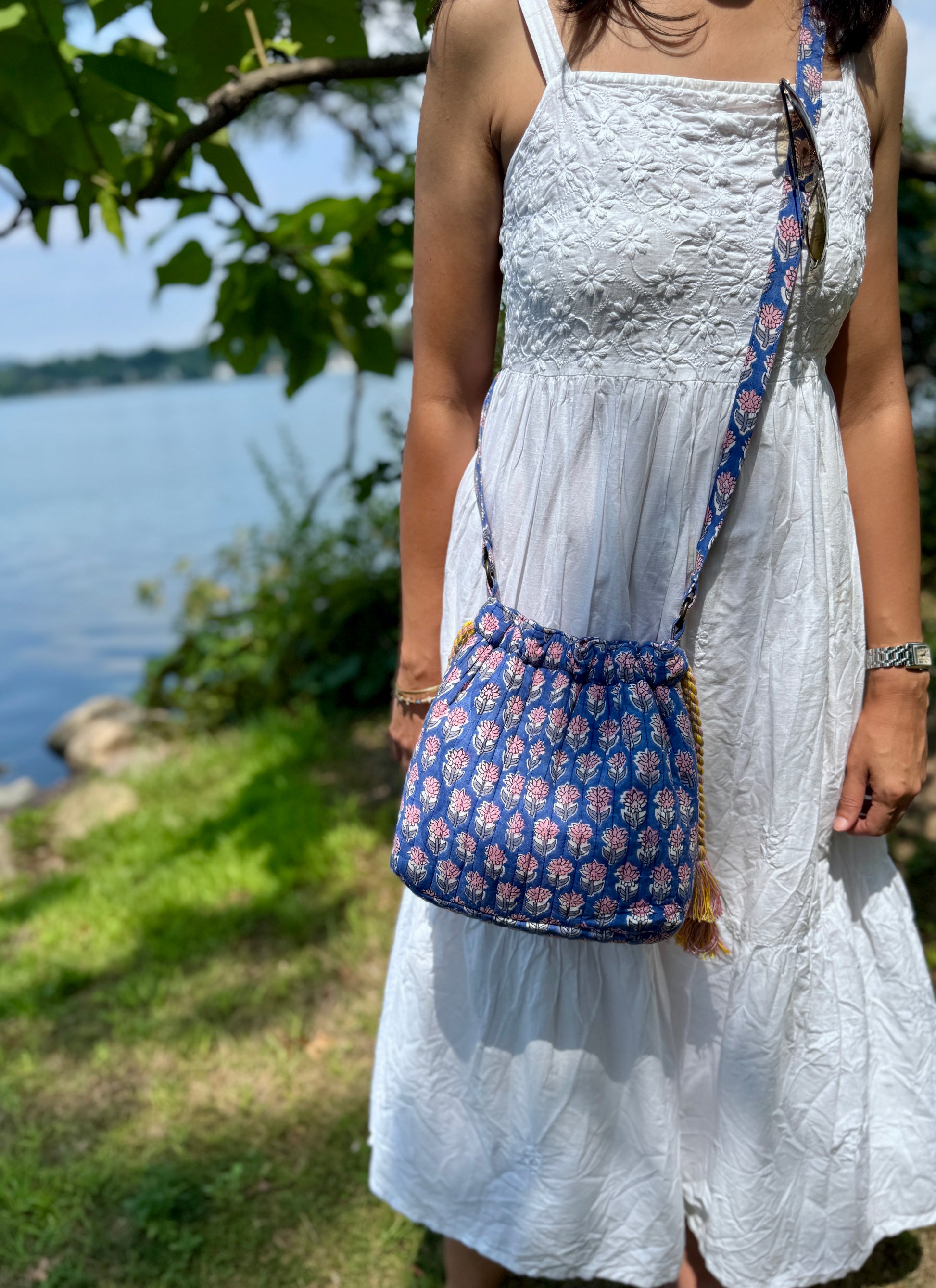 House of Mulmul - Garden Wayfarer Bucket Bag, Cotton bag, Crossbody sling bag. Woman in a white dress with a blue floral bag standing by a body of water.