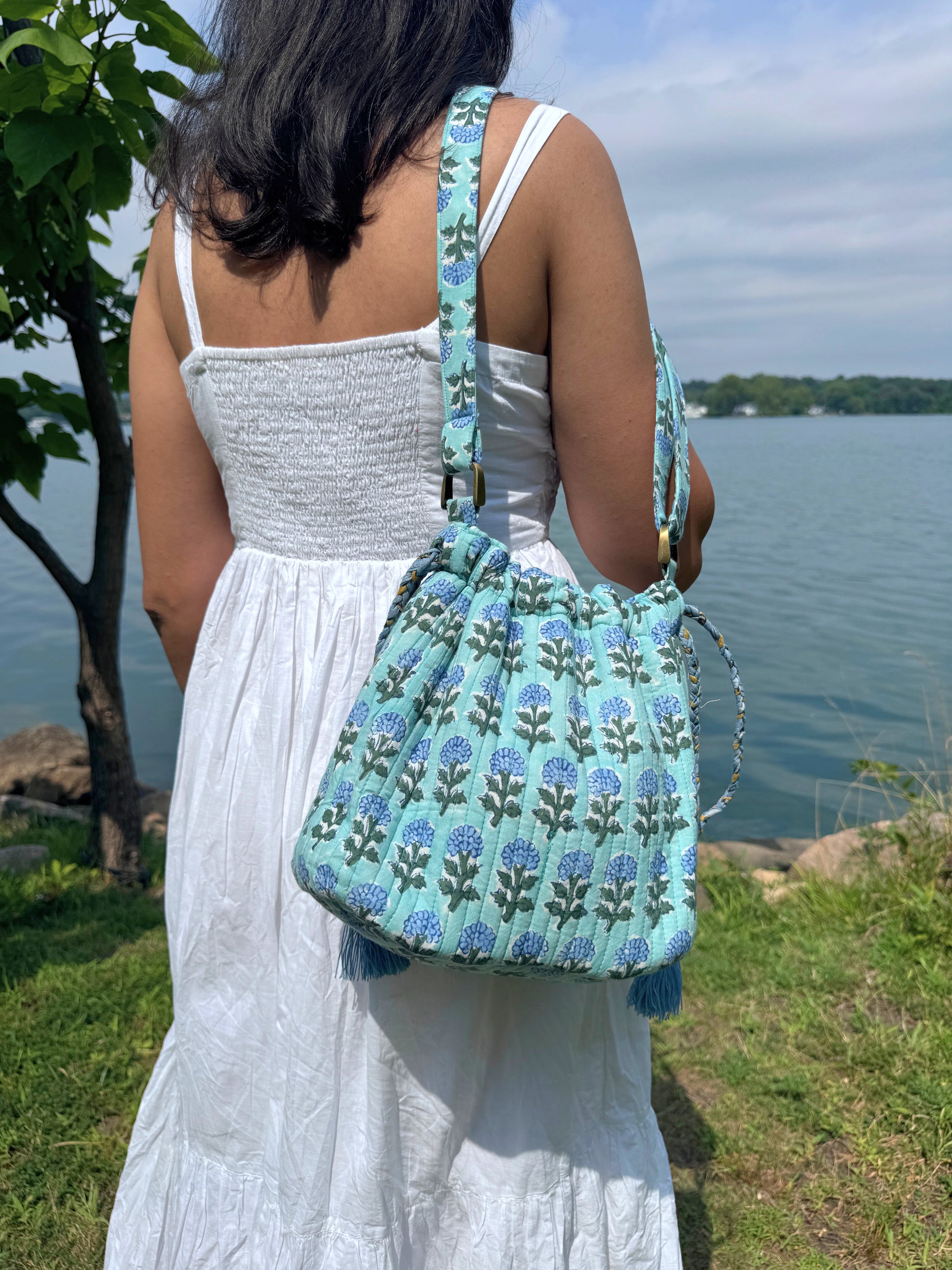 House of Mulmul - Garden Wayfarer Bucket Bag, Cotton bag, Crossbody sling bag. Person holding a blue floral-patterned bag by a body of water